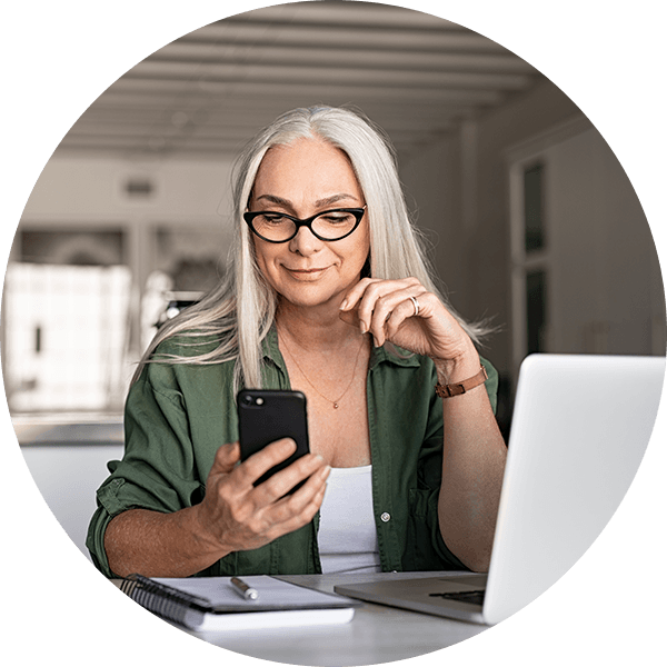 Older woman with white hair looking at smart phone while laptop and notebook is open.